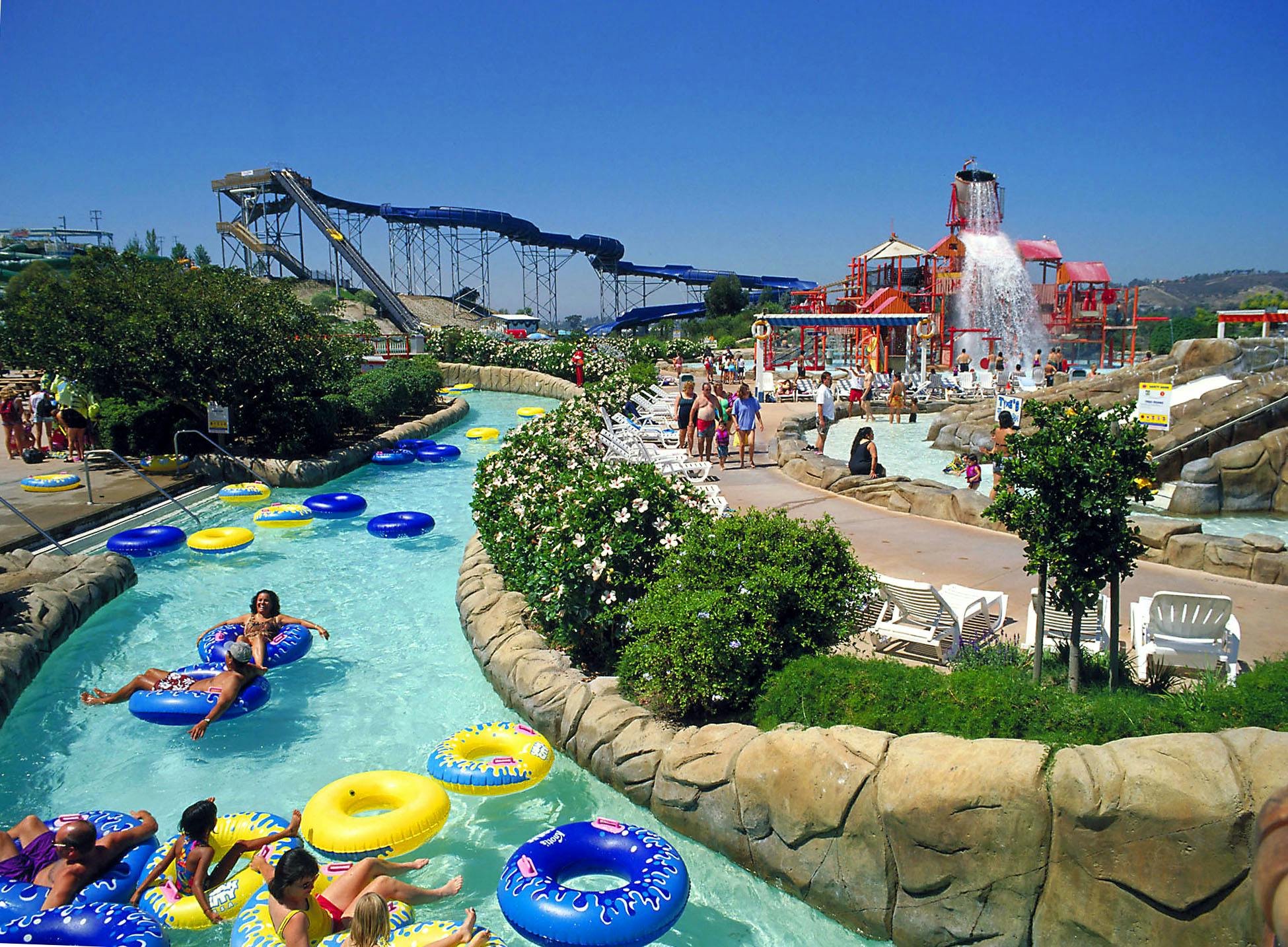Des personnes flottent dans une piscine sur des boudins gonflables près d'un bâtiment au décor nautique et de palmiers sous un ciel bleu.