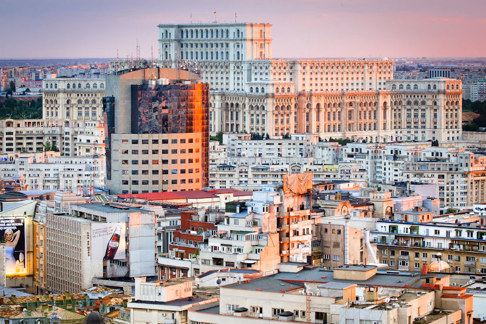 Aerial view of a densely packed urban area with various buildings, featuring a large, ornate palace in the background.