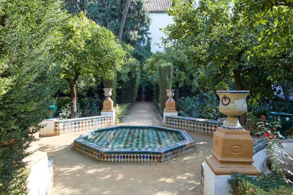 Ornate octagonal fountain with patterned tiles in a lush garden with trees, bushes, and two decorative urns on pedestals. Pathway leads ahead.
