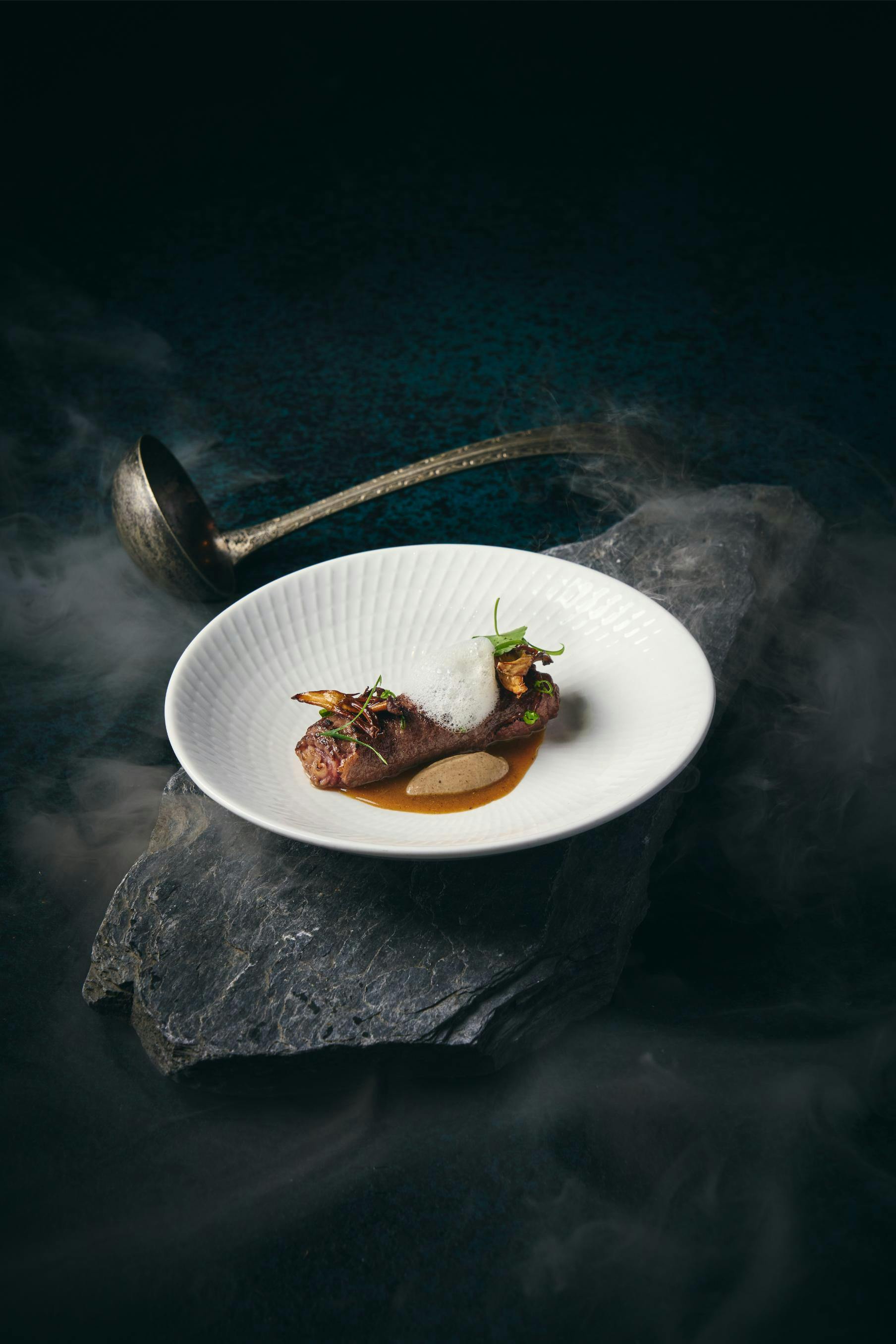 A gourmet dish plated on a white bowl placed on a stone slab, with a metal ladle in the background.