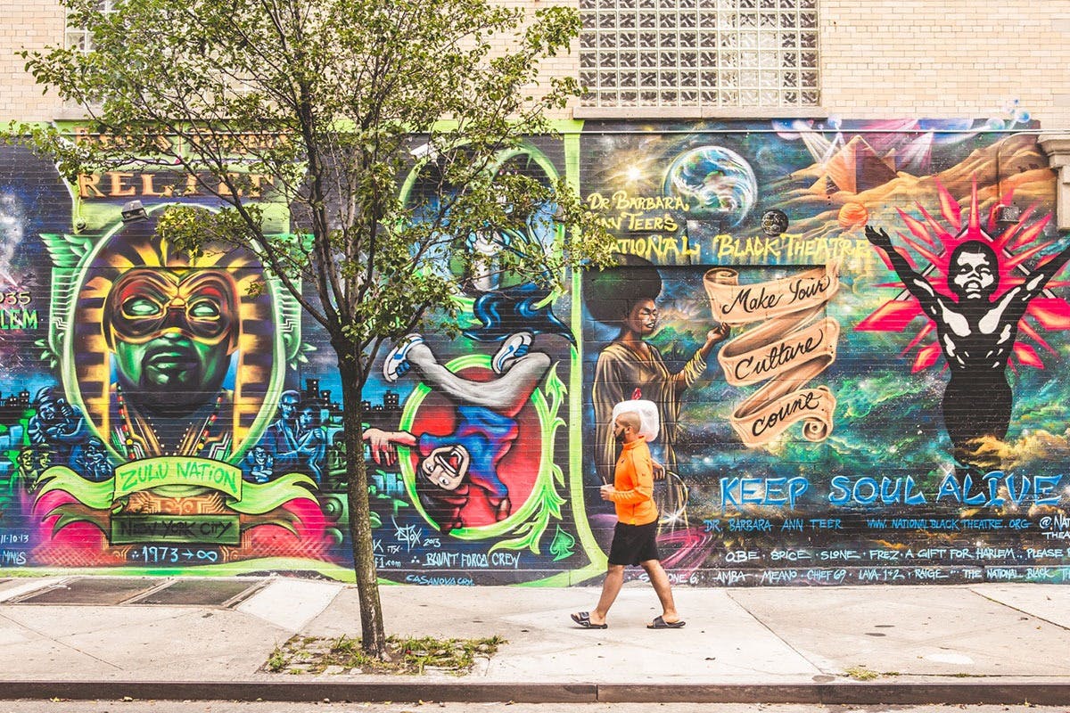 Человек в оранжевой рубашке проходит мимо красочной фрески с различными фигурами, смелым текстом и яркими космическими произведениями искусства.