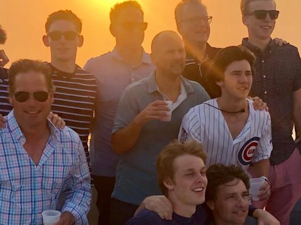A group of 14 people pose for a photo on a boat at sunset, with a calm sea in the background.