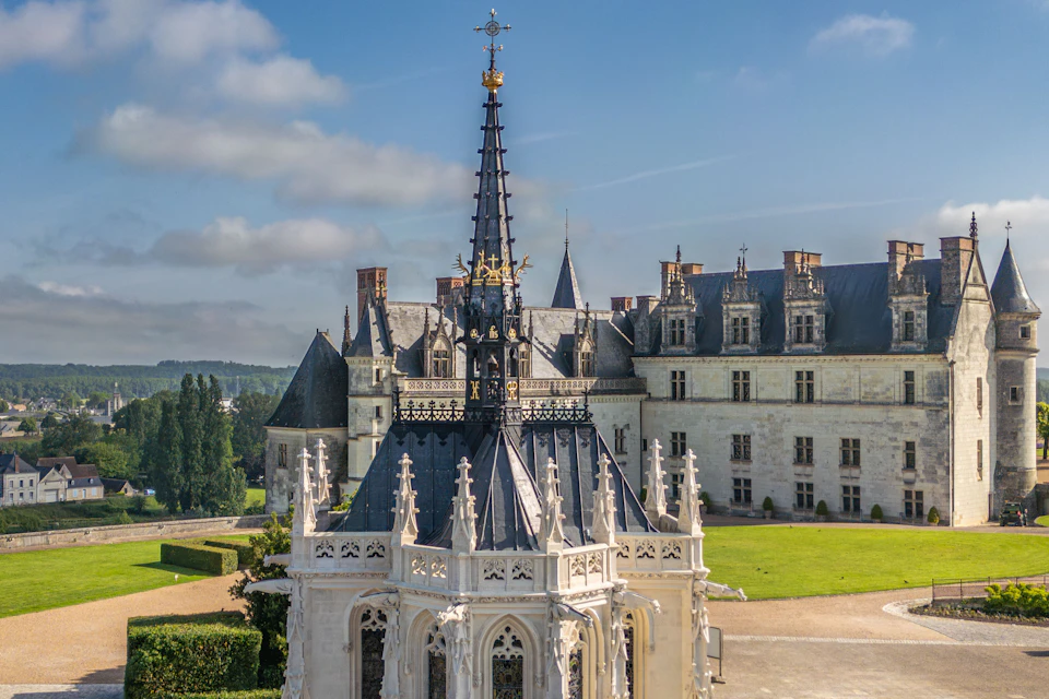 A gothic-style chapel with intricate carvings and a tall spire, set against a historic castle and a landscaped garden.