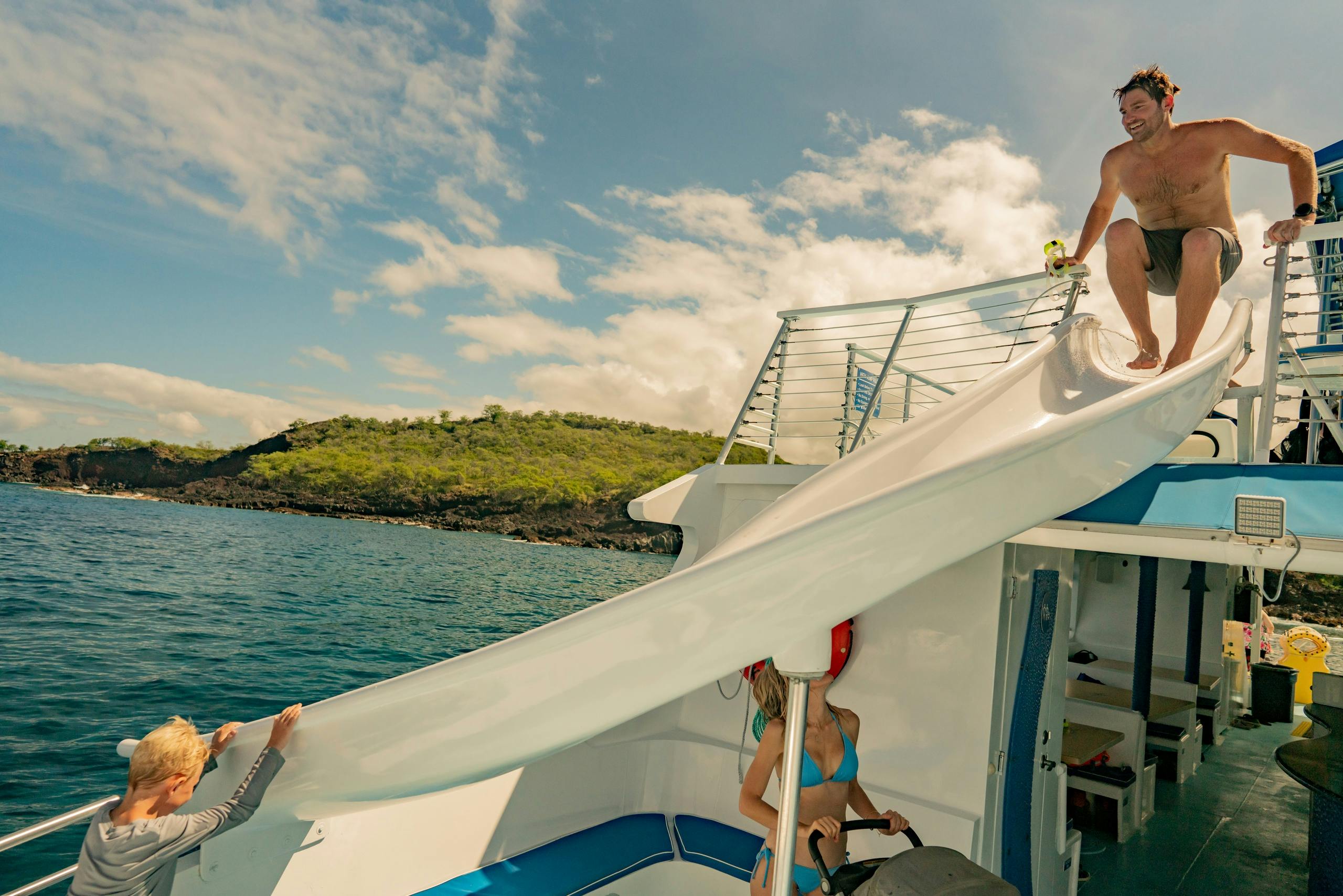 A man is about to slide down a waterslide on a boat, with a woman standing below in a swimsuit. The setting is outdoors near water.