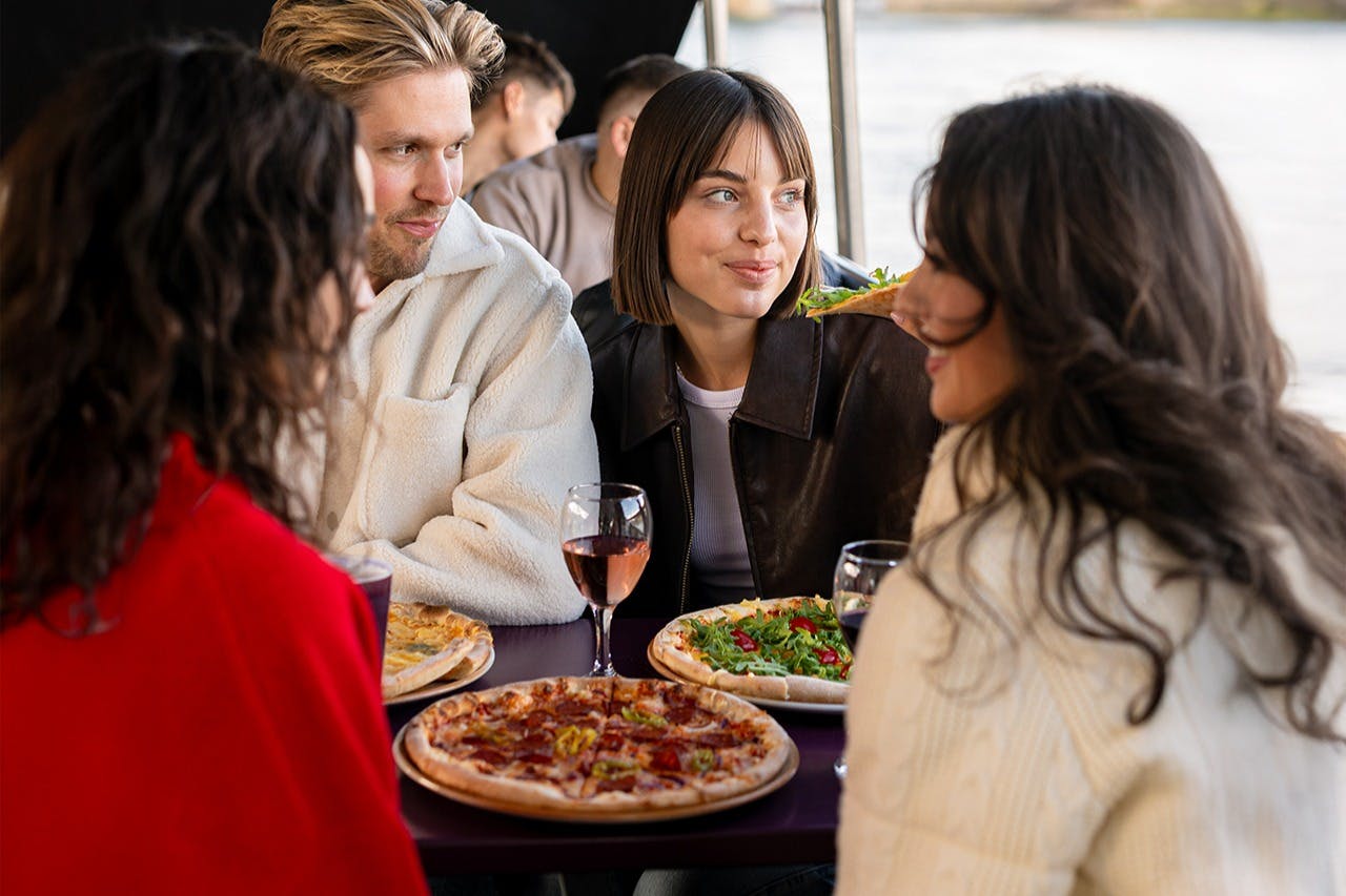 4명이 테이블에 앉아 다양한 피자와 와인잔을 마시며 미소를 짓고 대화에 참여합니다.