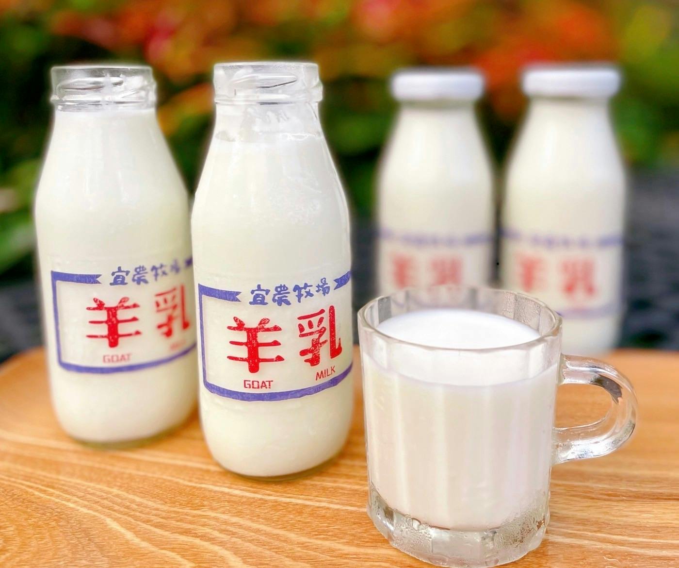 Four labeled bottles of goat milk with a clear mug of milk, set on a wooden surface with blurred greenery in the background.