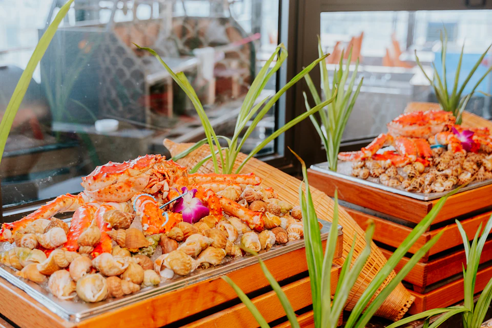 A wooden platter filled with seafood, including crab legs and shells, decorated with plants, placed by a window.