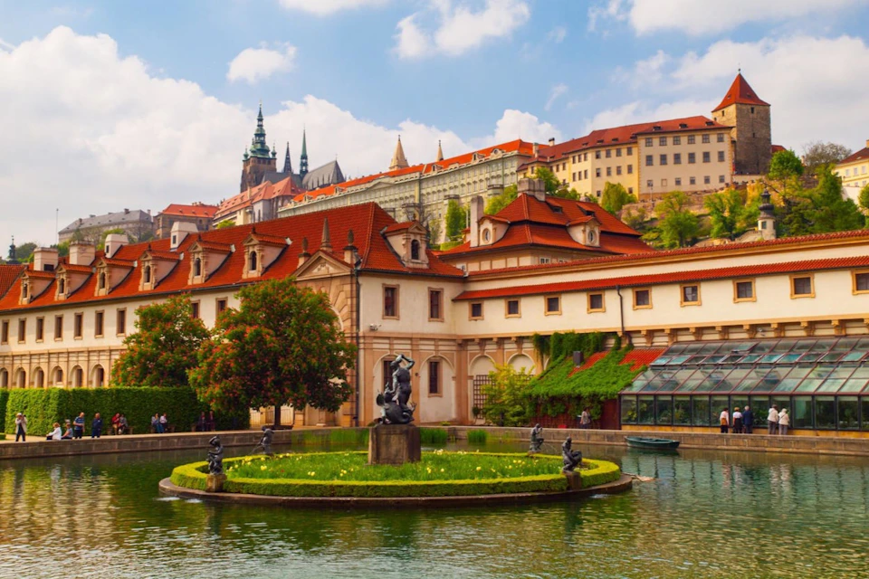 A scenic view of a historical building with red roofs, a garden, a fountain, and statues in the foreground under a partly cloudy sky.