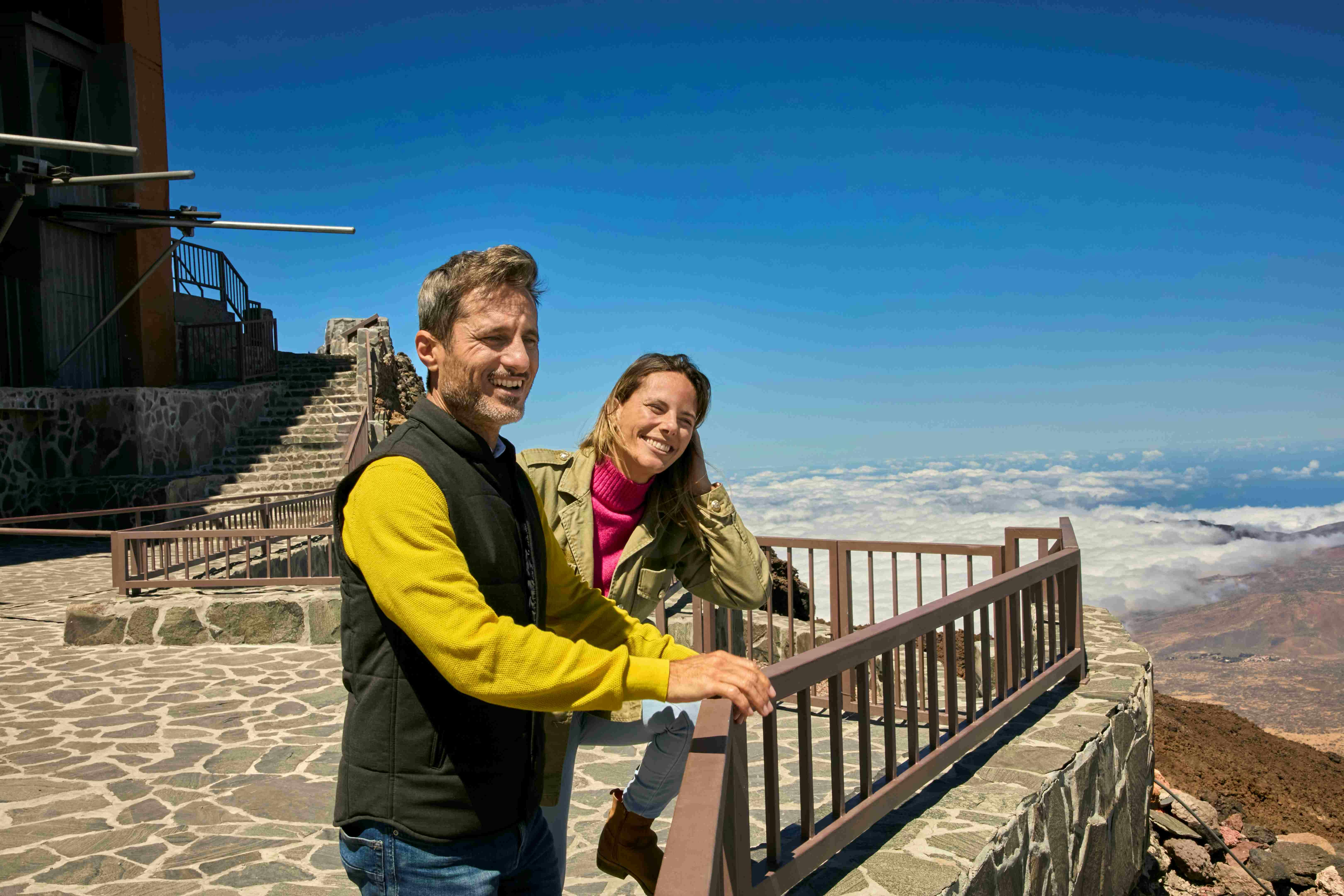 Dues persones somrient recolzades en una barana d'un mirador, amb una passarel·la de pedra i escales. Núvols a sota i cel blau a sobre.