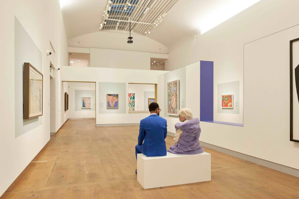 Two people sit on a bench in an art gallery, observing colorful paintings displayed on the walls under bright lighting.