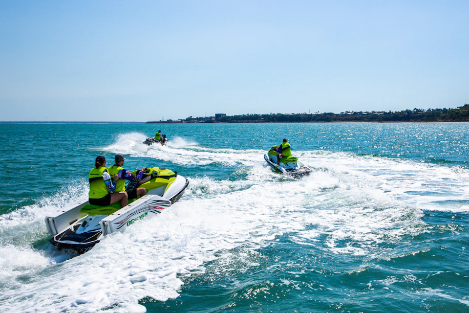 People riding jet skis in turquoise water on a sunny day, with land visible in the background.