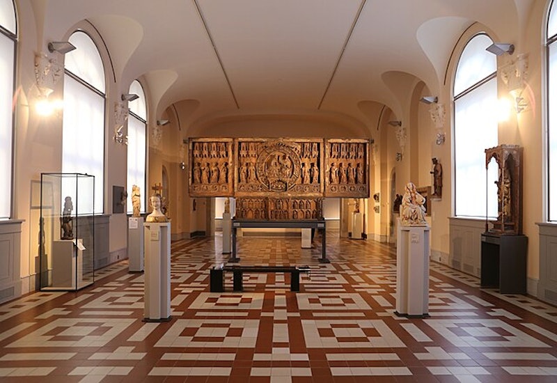 A spacious museum gallery with statues on pedestals, intricate wooden art on the back wall, and a geometric tiled floor.