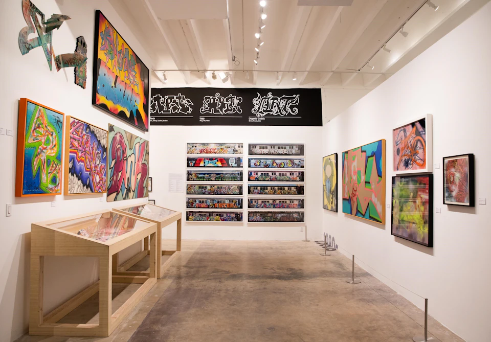 A gallery with vibrant graffiti art displayed on walls and in glass cases, under bright ceiling lights.