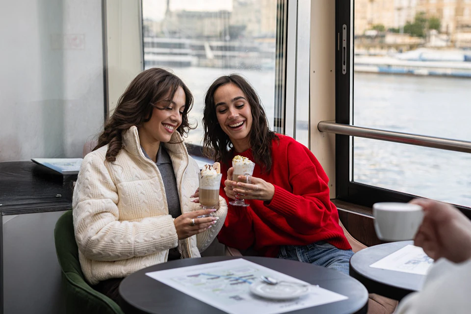 Two women sitting by a window, smiling and holding drinks topped with whipped cream at a cafe table.