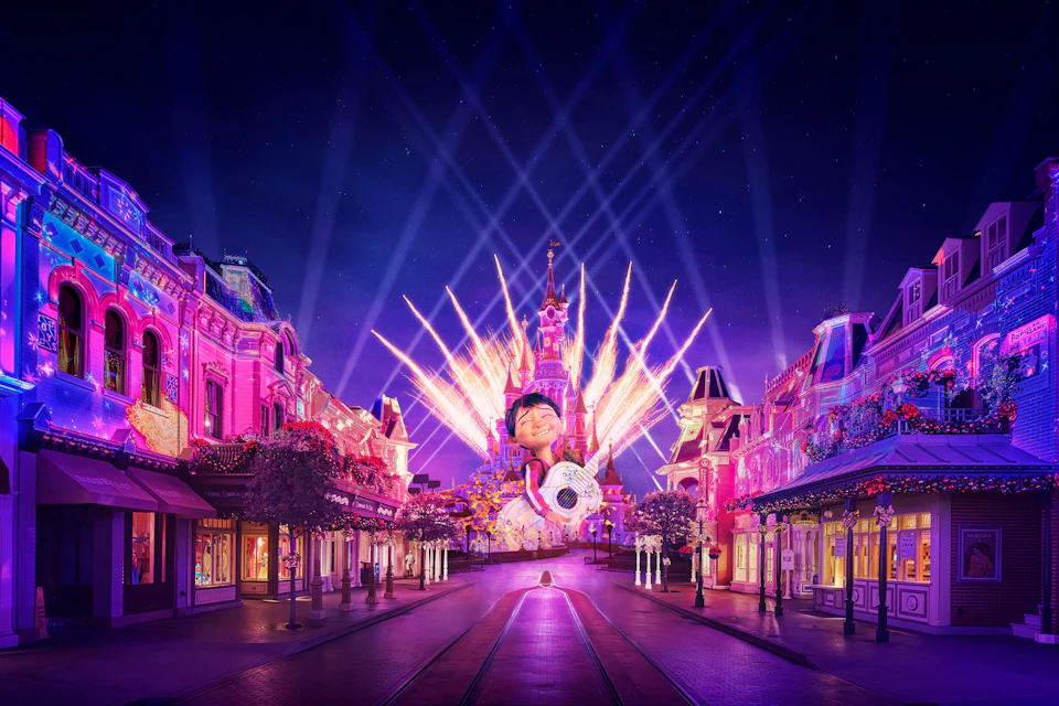 Colorful fireworks illuminate a castle at night, with a wide street leading towards it. Buildings on either side are lit in pink.