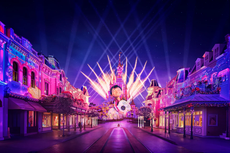 Colorful fireworks illuminate a castle at night, with a wide street leading towards it. Buildings on either side are lit in pink.