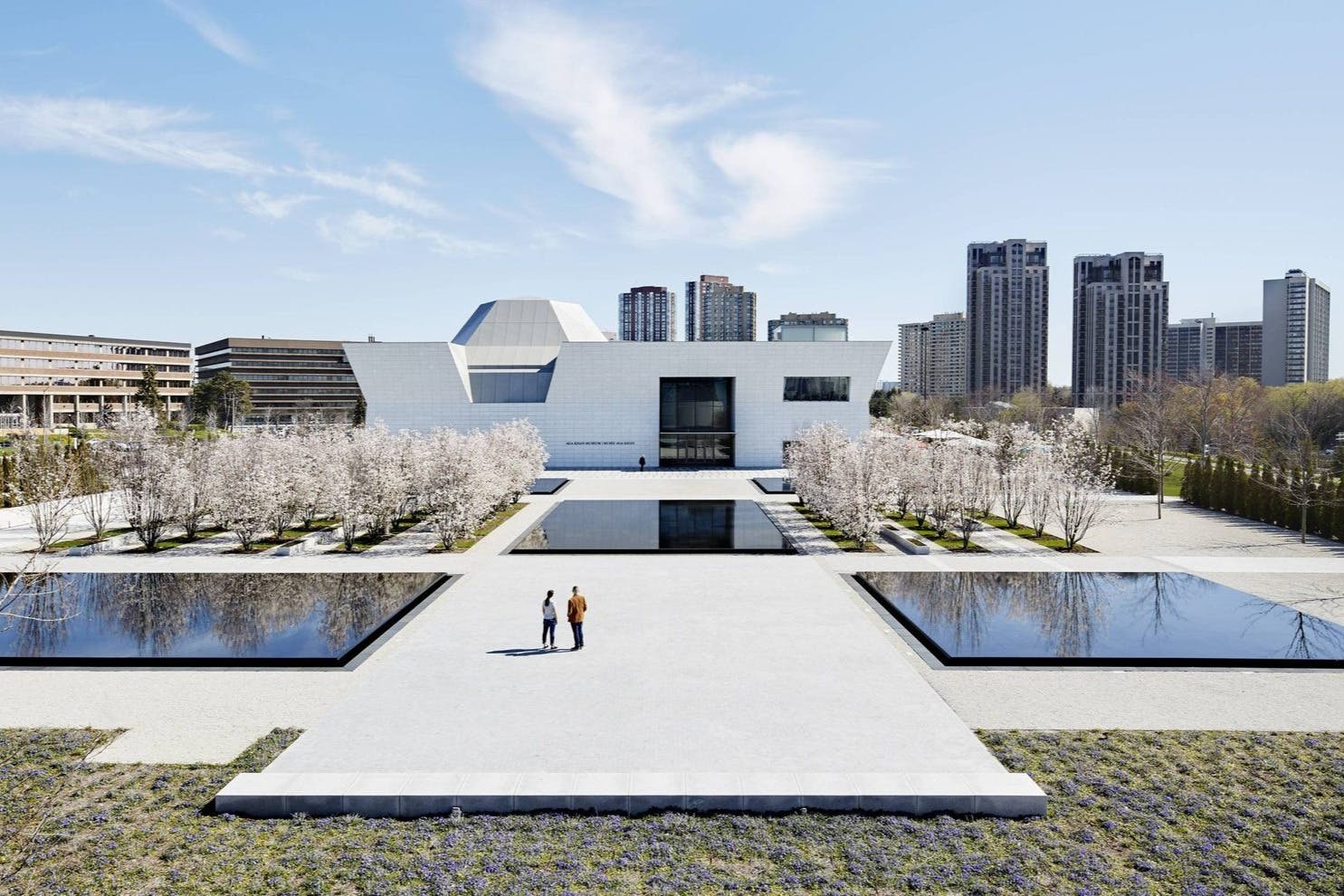 Moderní bílá budova s reflexními bazény, obklopená kvetoucími stromy. Dva lidé jdou ke vchodu pod jasnou oblohou.