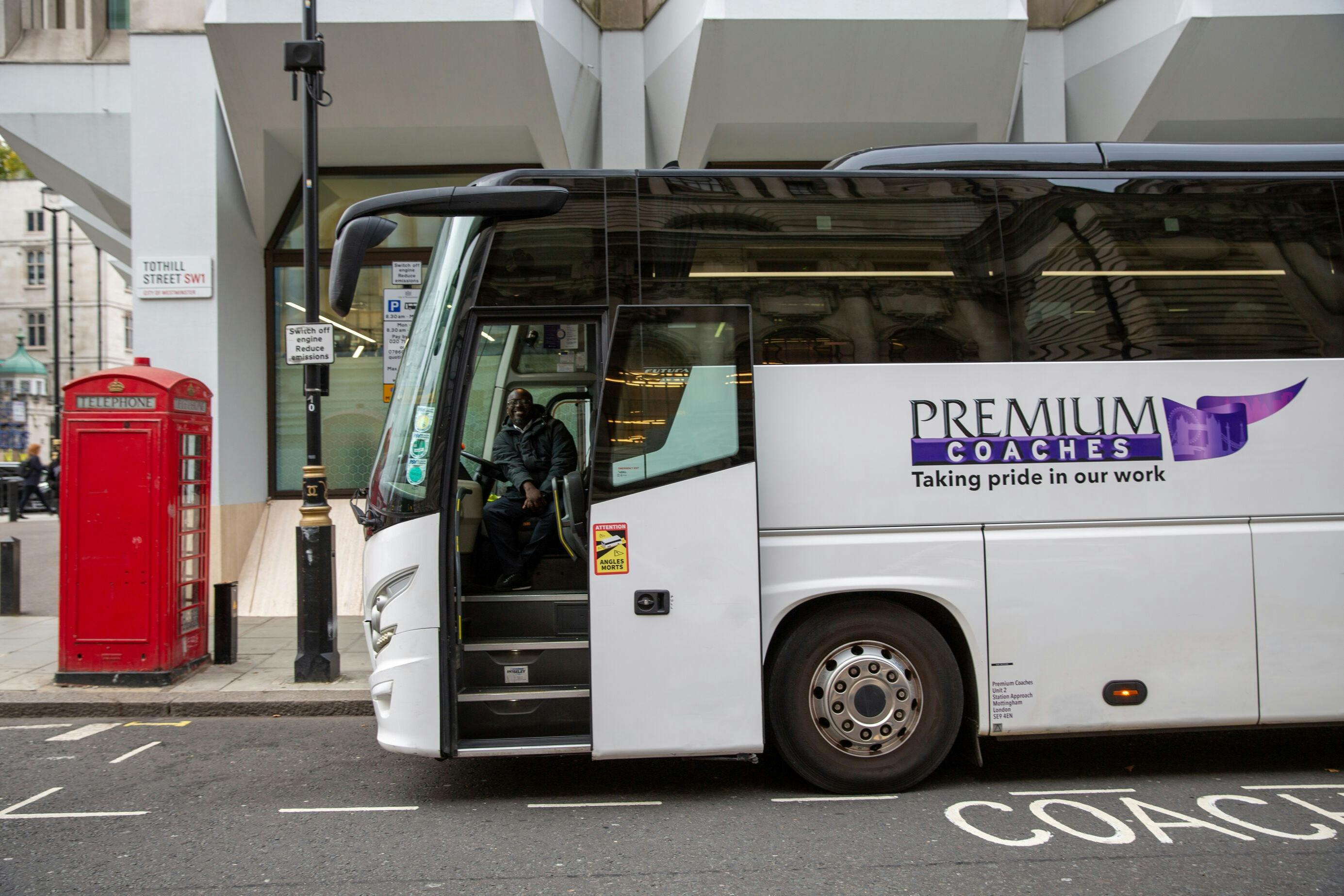 En hvid bus mærket "Premium Coaches" parkerede på en gade med døren åben og en person synlig indeni.