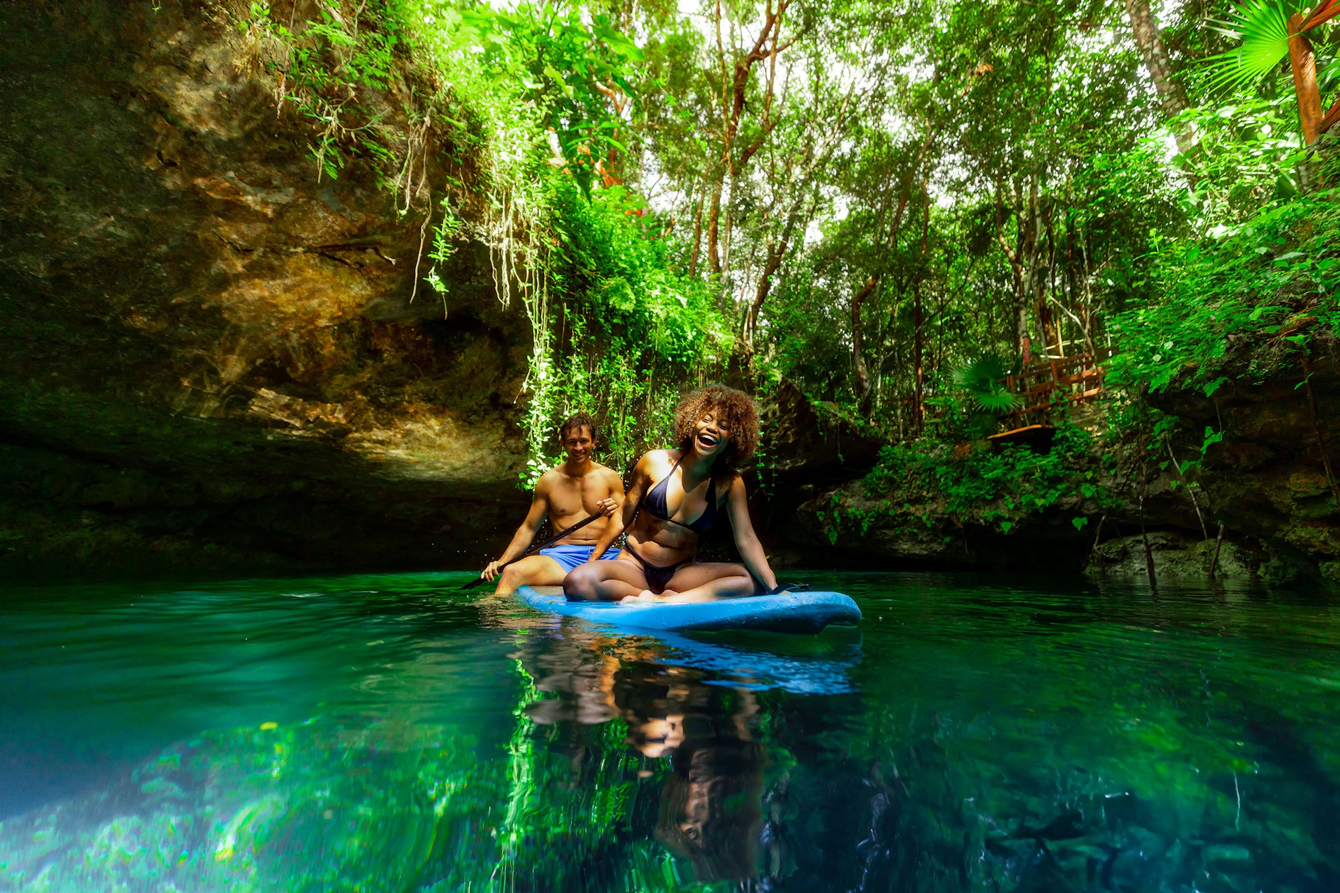 Two people enjoy paddleboarding on clear water surrounded by lush greenery and rocky cliffs in a natural setting.