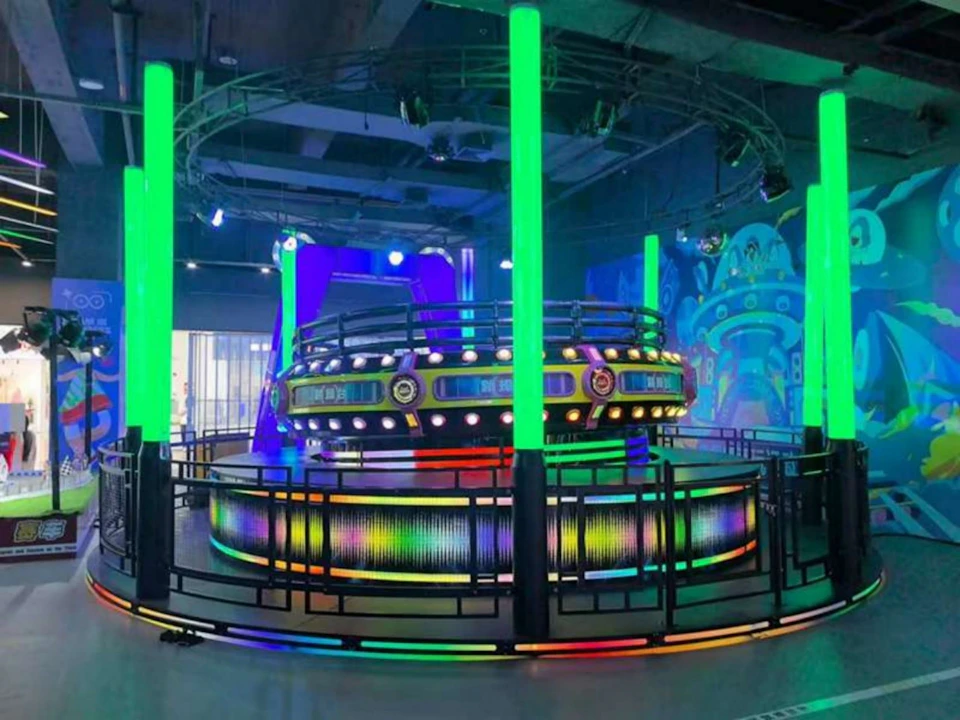 Indoor amusement ride with neon green and multicolor lights, surrounded by railings and a vibrant futuristic theme.