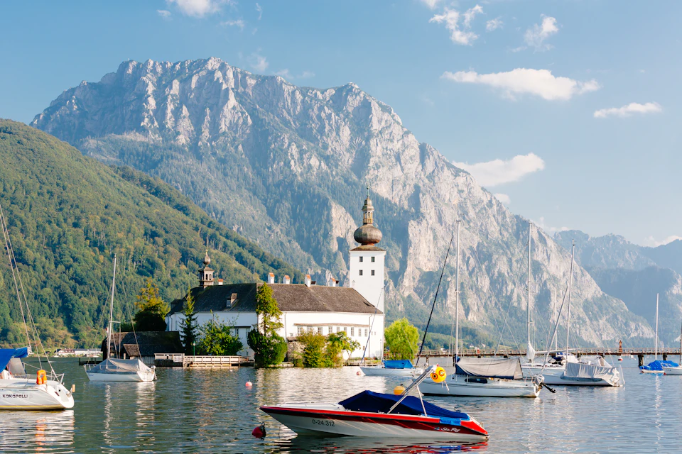 A lakeside scene with boats, a white church with a dome, and mountains in the background under a partly cloudy sky.