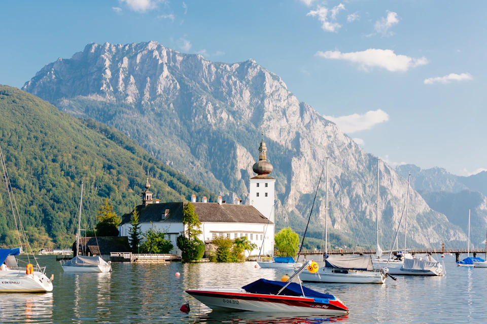 A lakeside scene with boats, a white church with a dome, and mountains in the background under a partly cloudy sky.