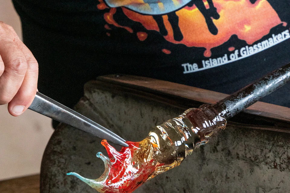 A close-up of a glassblowing process, with molten glass being shaped using tools, against a colorful background.
