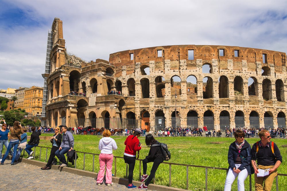 Tourists at the Colosseum in Rome on a cloudy day, with people walking and standing near the ancient amphitheater.