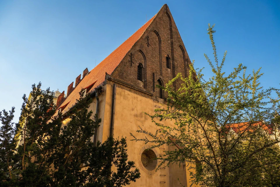 A historic building with a steep triangular roof, arched windows, and trees partially obscuring the facade, under a clear sky.