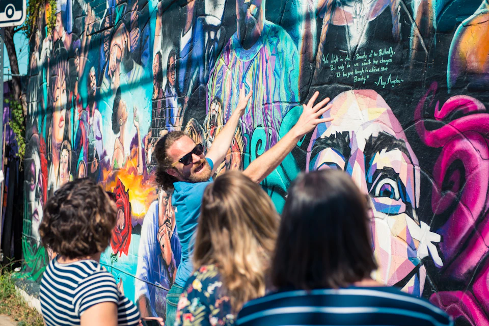 A person in sunglasses gestures towards a colorful mural while a group of people listens attentively.