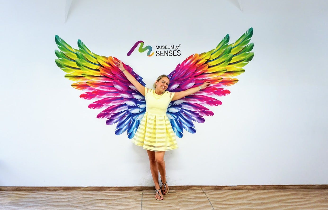 A woman in a yellow dress poses with outstretched arms in front of a colorful, wing-shaped mural at the Museum of Senses.