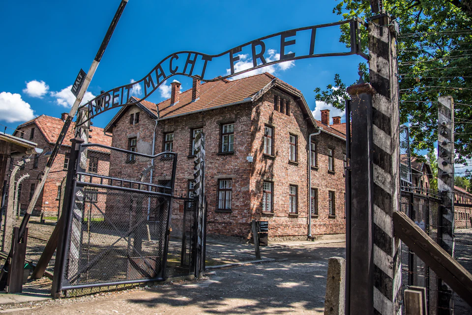 Brick buildings behind a gate with the inscription "Arbeit Macht Frei" beneath a blue sky with clouds.