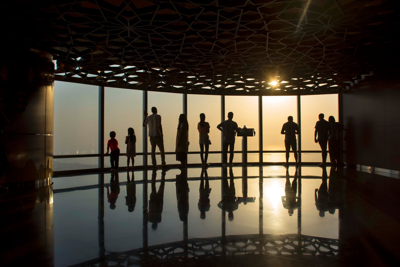 Silhouetted people stand in front of large windows during sunset on a glossy floor under an intricate patterned ceiling.