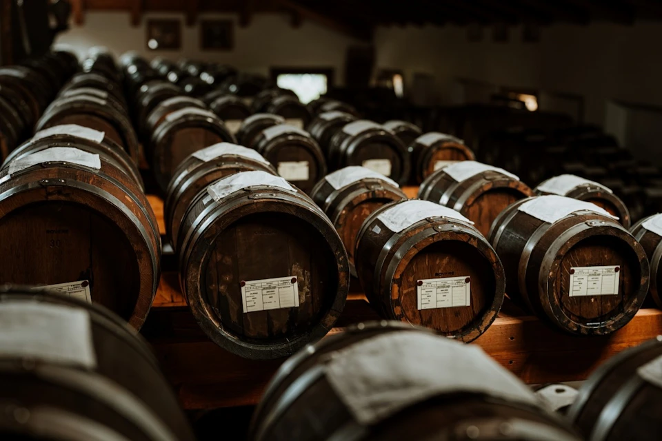 Rows of stacked wooden barrels with labels in a dimly lit storage room.