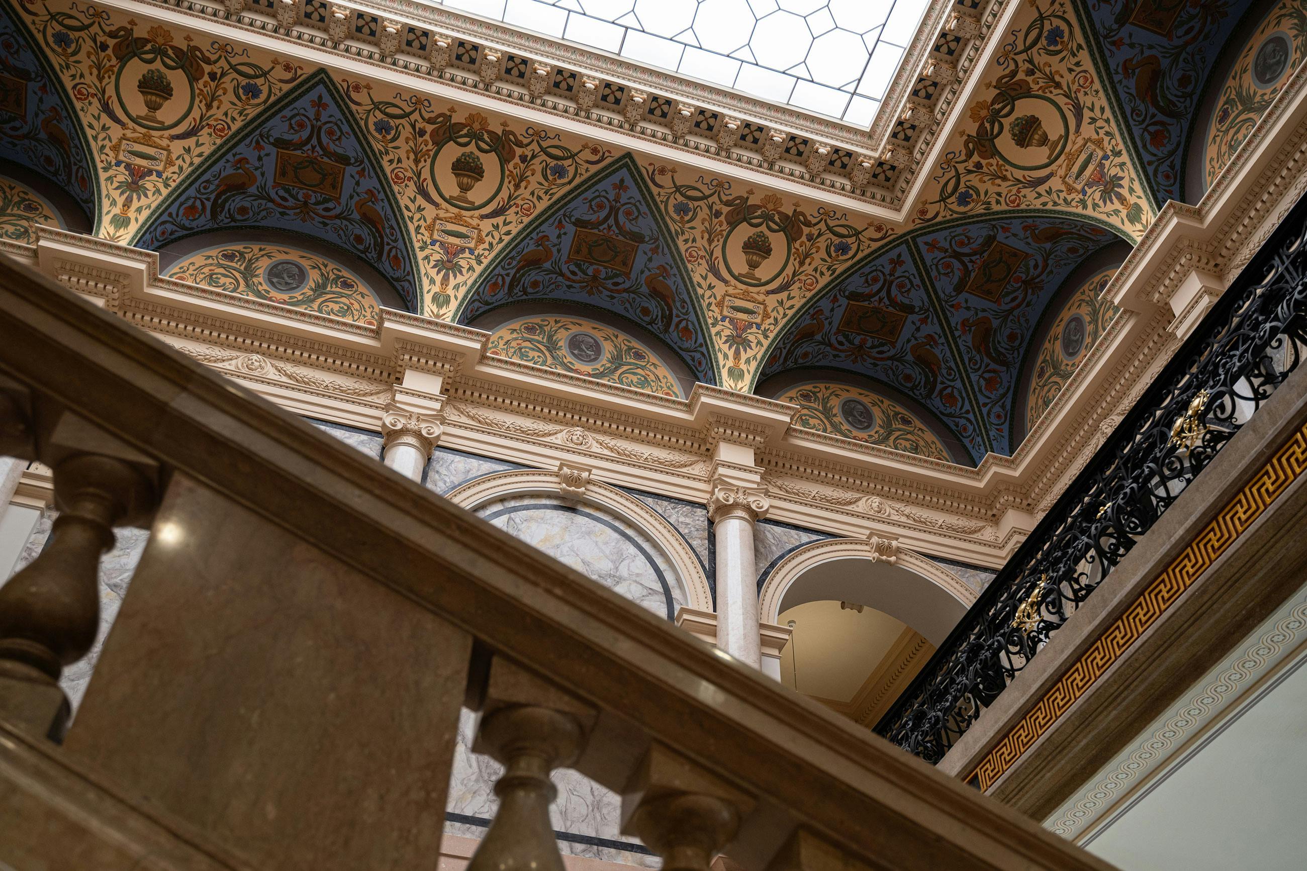 Περίτεχνο εσωτερικό με διακοσμημένη οροφή, τοξωτές κόγχες τοίχων, κολώνες και μαρμάρινο κιγκλίδωμα σκάλας στο προσκήνιο.