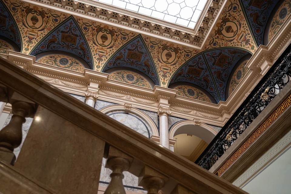 Ornate interior with a decorated ceiling, arched wall niches, columns, and a marble staircase railing in the foreground.