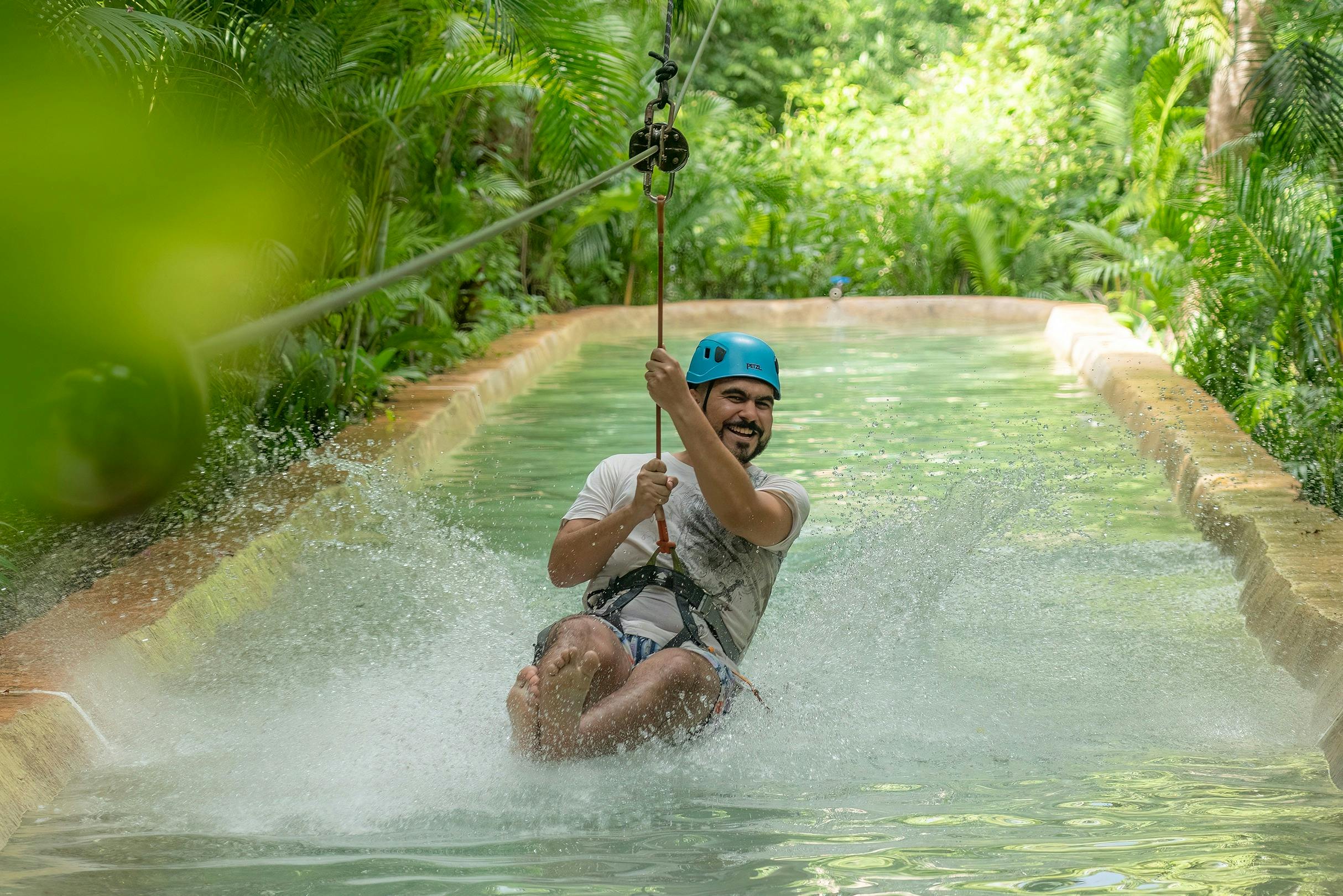 Un home que porta un casc blau fa tirolesa sobre un curs d'aigua, creant esquitxades i envoltat de vegetació exuberant.