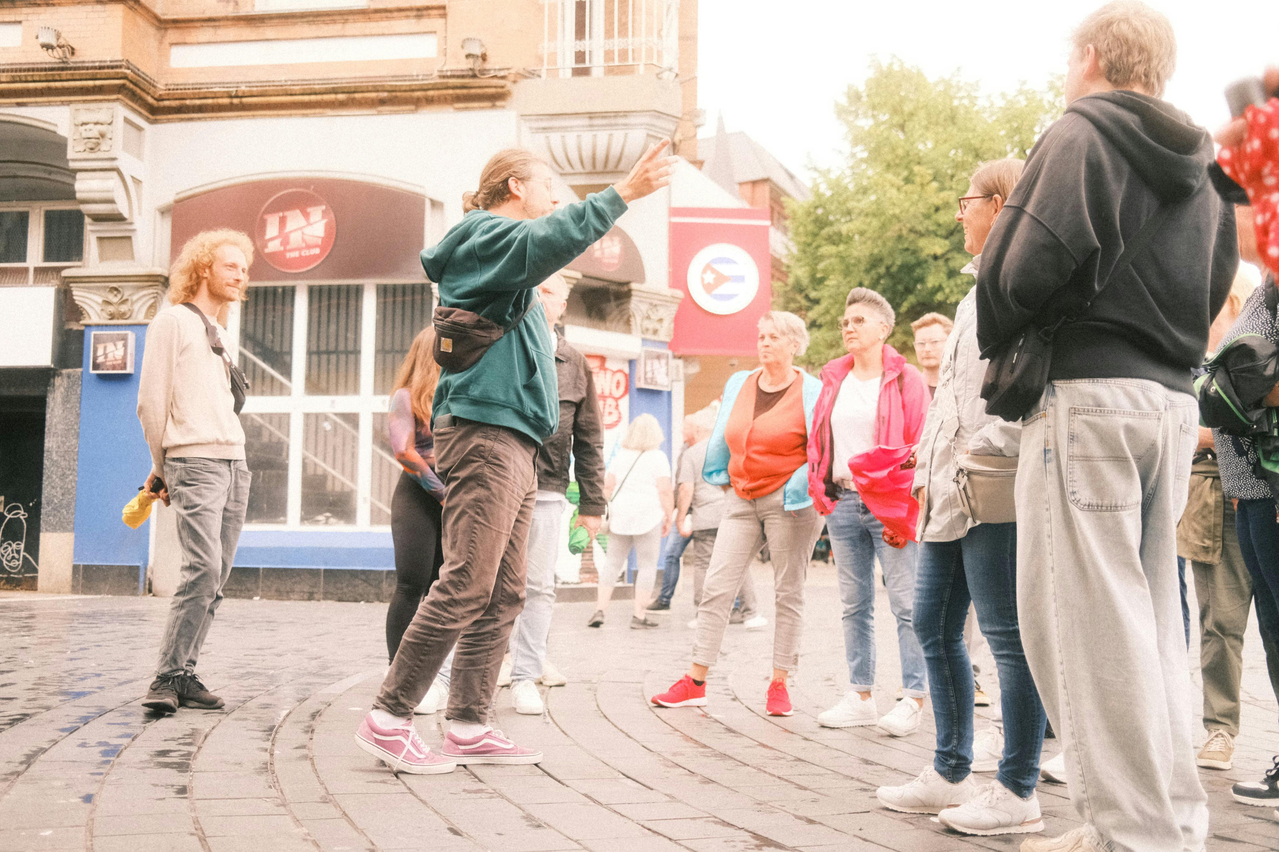 Ένας ξεναγός κάνει χειρονομίες ενώ μιλάει σε μια ποικιλόμορφη ομάδα ανθρώπων σε μια πλατεία της πόλης. Τα κτίρια και το πράσινο βρίσκονται στο παρασκήνιο.