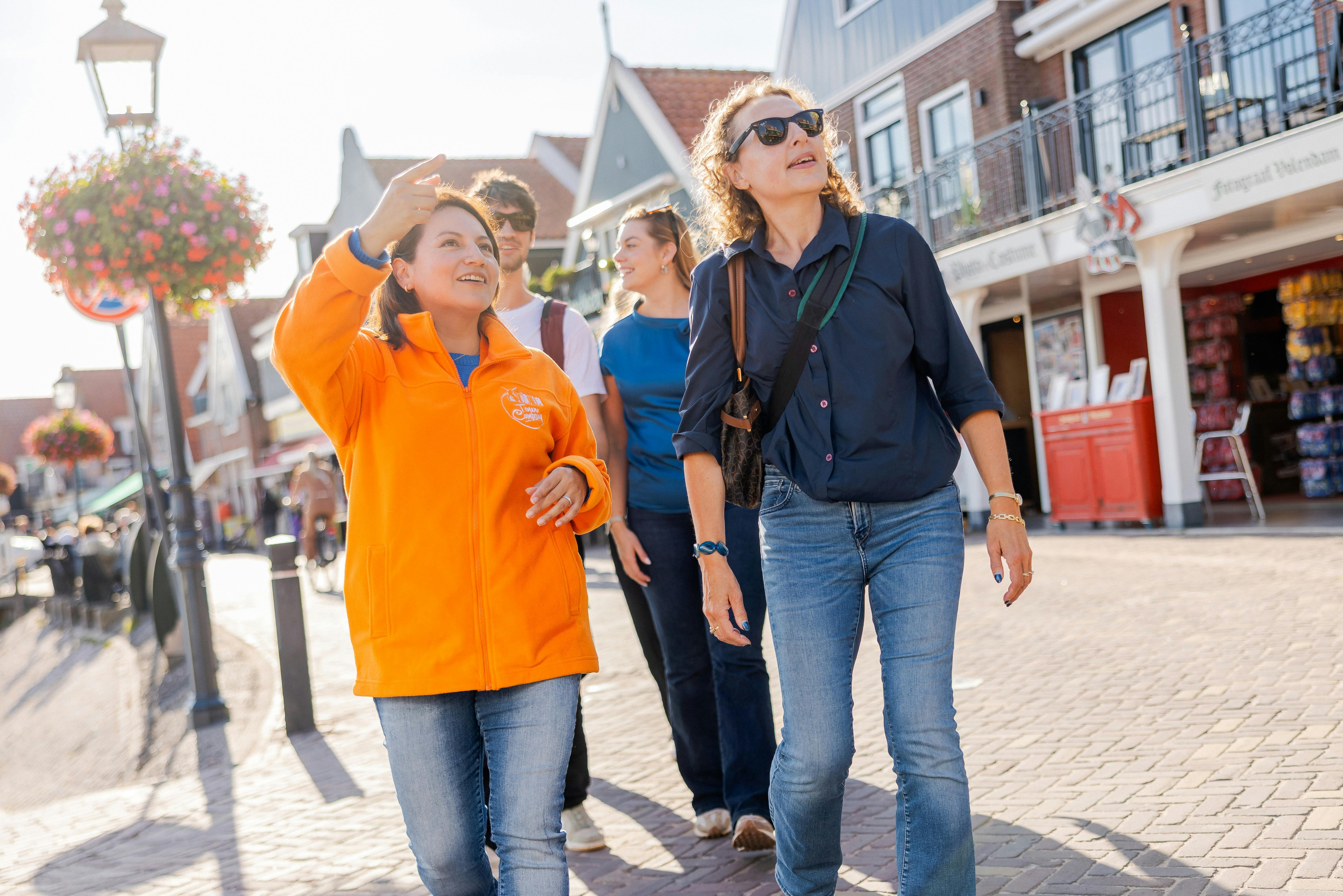 A woman in an orange jacket gestures as she leads a group of people through a sunny, cobblestone street with shops and houses.