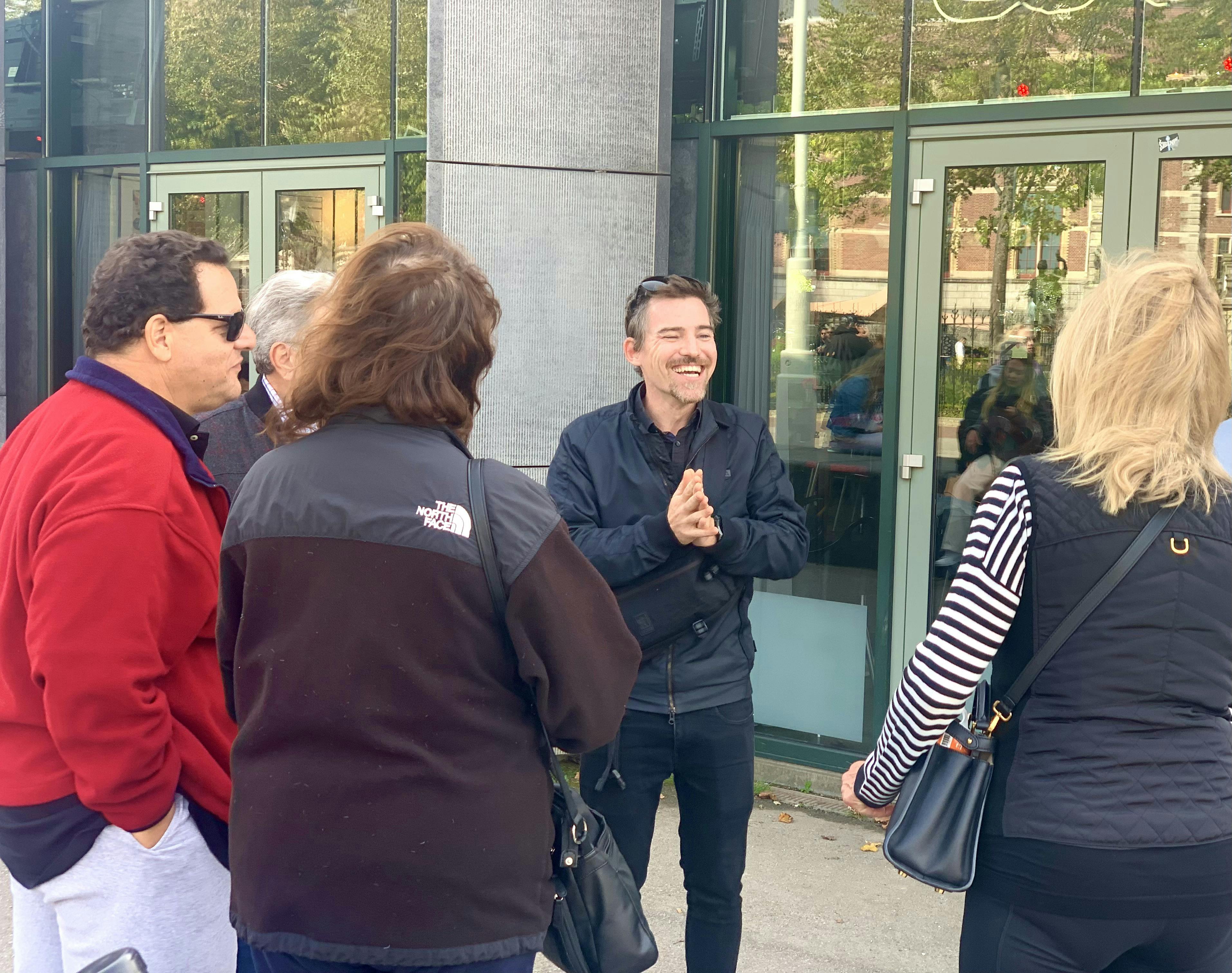 一名男子微笑着拍手，同时在玻璃门窗的建筑物外与一小群人交谈。