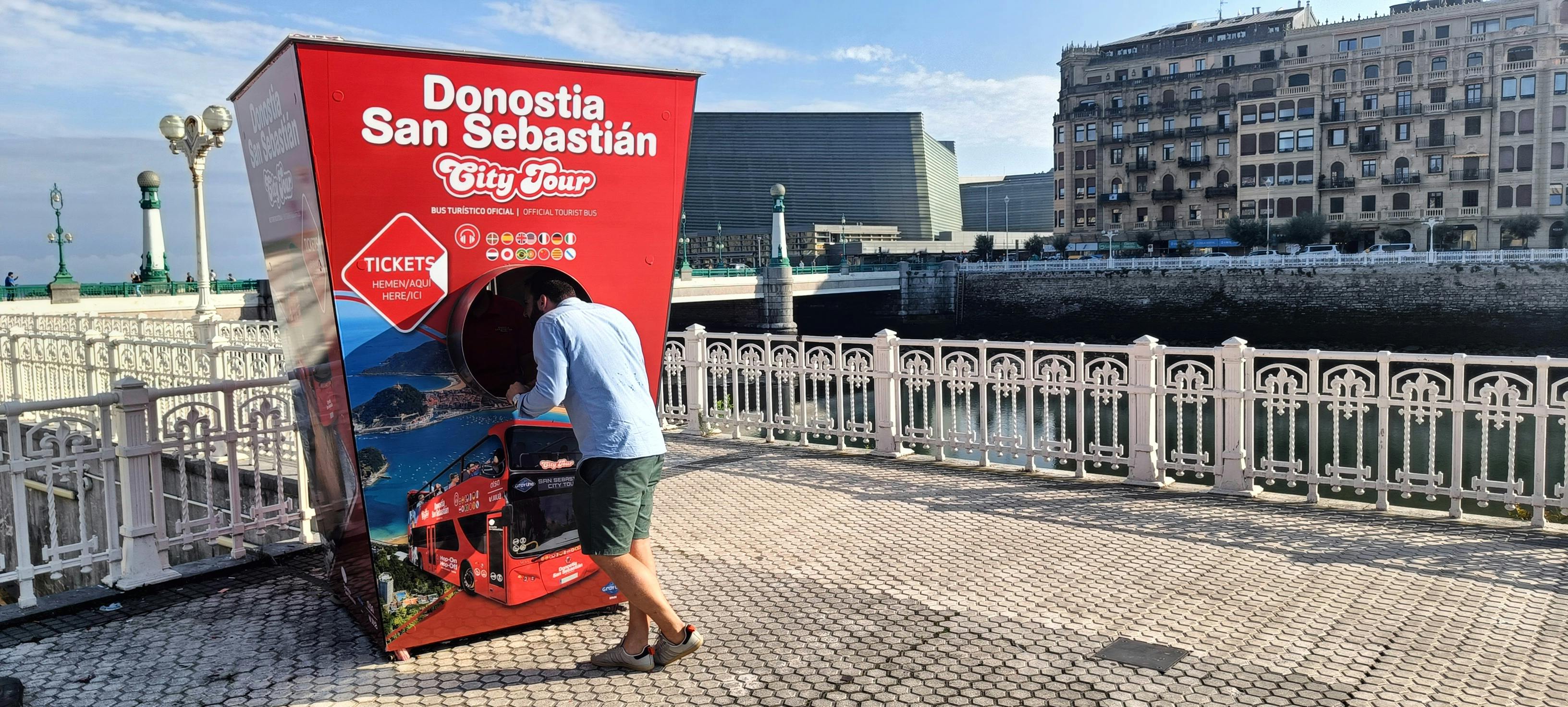 A red double-decker tour bus travels past a historic stone building with arched windows under a clear blue sky.