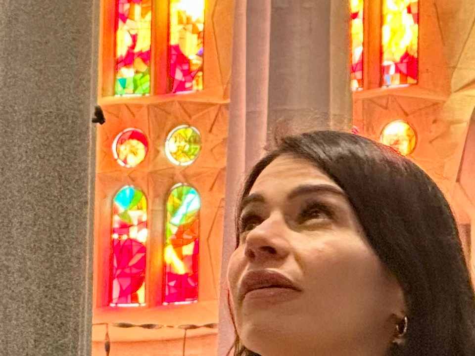 Two women inside a large, brightly lit building with colorful stained glass windows. One woman is pointing upwards.