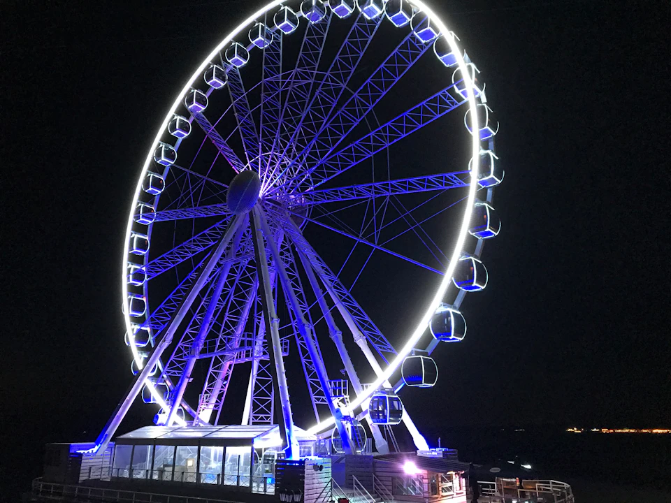 A brightly lit Ferris wheel with enclosed cabins illuminated in blue and white lights at night.
