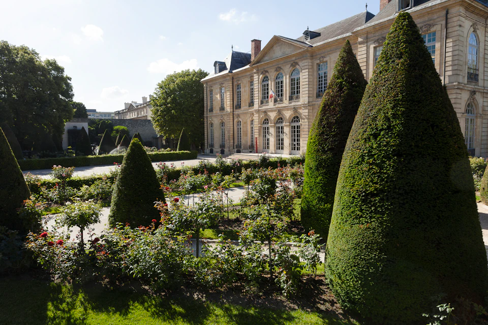 Elegant garden with manicured bushes and flowerbeds in front of a large, historic building with arched windows.