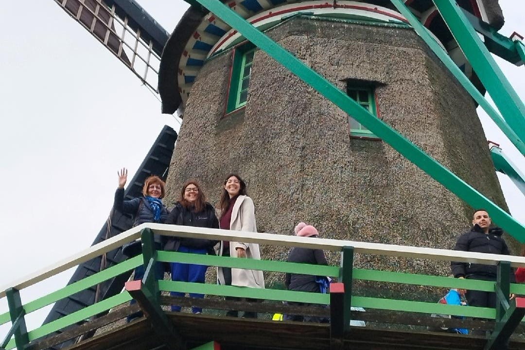 Tři lidé stojí na balkoně větrného mlýna, usmívají se do kamery a jeden mává. Obloha je zatažená.