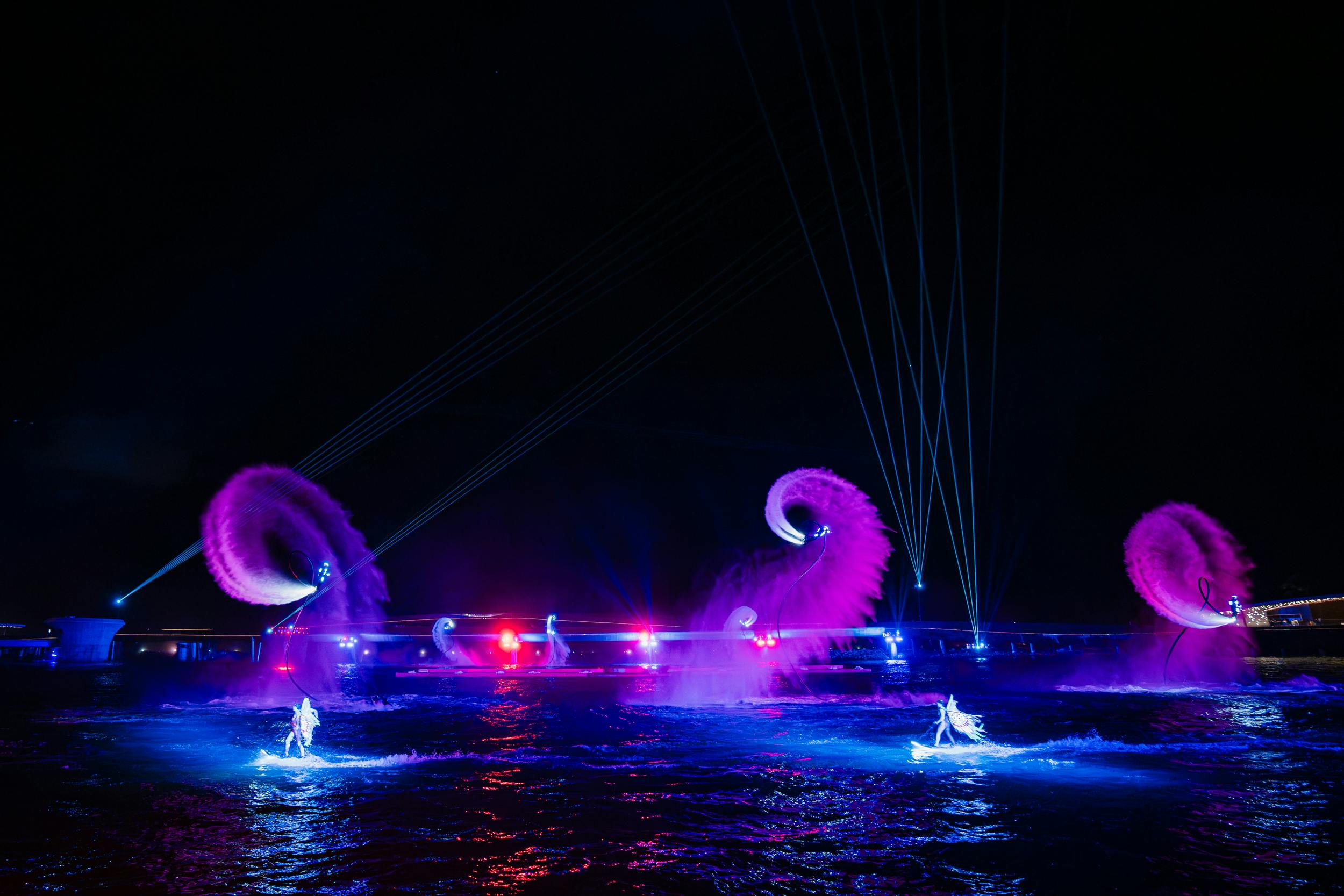 A nighttime water performance featuring colorful lights, water jets, and illuminated performers on a water surface.