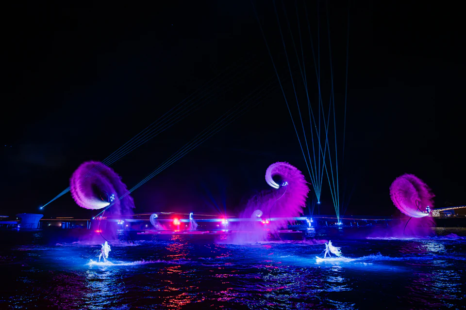 A vibrant night water show with illuminated performers, colored lights, and water sprays creating dynamic patterns.