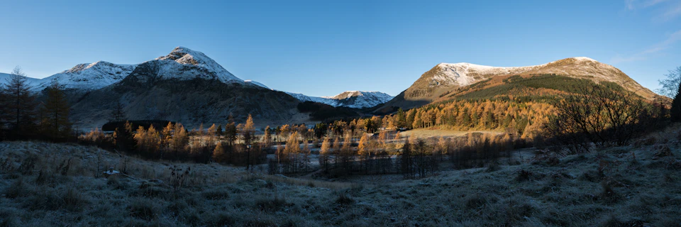 A vast, frosty valley with sparse trees and mountain backdrop, contrasted against a clear blue sky illuminated by soft sunlight.