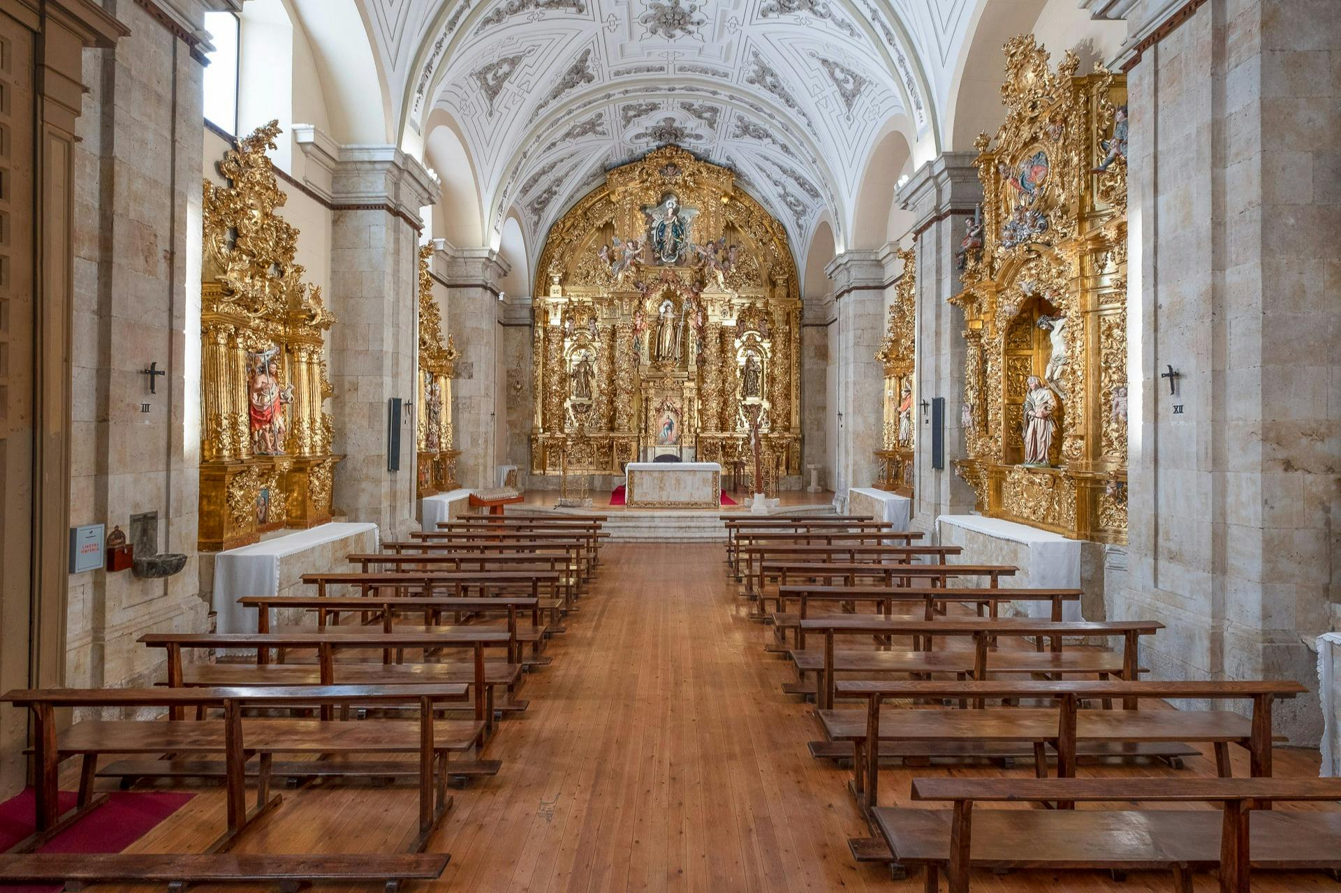 Un intérieur d'église orné de bancs en bois, d'autels dorés et de détails complexes au plafond, centré sur un autel décoré.