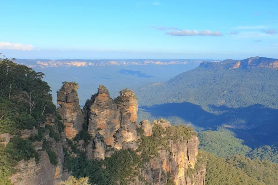 Three towering rock formations, known as the Three Sisters, surrounded by forested valleys and distant mountain ranges under a clear blue sky.